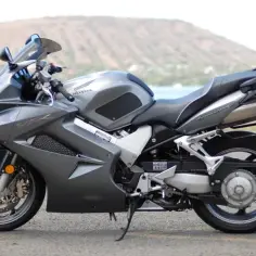 Szary motocykl Honda VFR 800 VTEC z widokiem na wodę i wzgórza w tle.