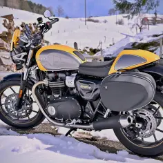 Żółto-szary motocykl Triumph Street Cup w zimowej scenerii. Silnik ma chłodzenie powietrzem, a sakwy boczne dodają mu uroku.