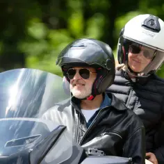 Uśmiechnięty mężczyzna w kasku i okularach przeciwsłonecznych, z pasażerką na motocyklu. Idealny wybór, jaki motocykl dla 50-latka?