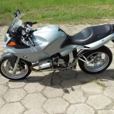 Srebrny motocykl BMW R1100S na betonowym placu, gotowy do jazdy po zielonych terenach.