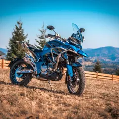 Niebieski motocykl turystyczny na trawiastym wzgórzu, z widokiem na góry i las.