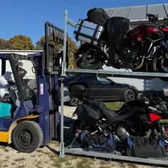 Wózek widłowy przygotowuje transport motocykla. Dwa motocykle na stojaku, jeden czerwony, drugi czarny.