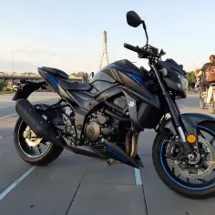 Czarny motocykl Suzuki GSX 750 na tle mostu. W tle ludzie spacerują i jeżdżą na rowerach.