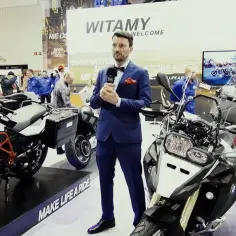Mężczyzna w garniturze prezentuje motocykl BMW 800 na targach. Obok stoją inne maszyny BMW.
