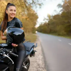 Uśmiechnięta kobieta w skórzanym stroju trzyma kask obok swojego motocykla. Idealne motocykle dla kobiet czekają na przygodę.