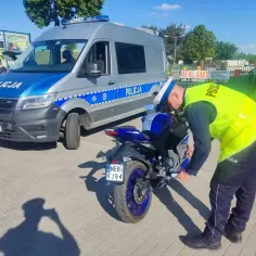 Policjant sprawdza dokumenty motocyklisty, by upewnić się, czy przelot jest legalny. Obok stoi radiowóz.