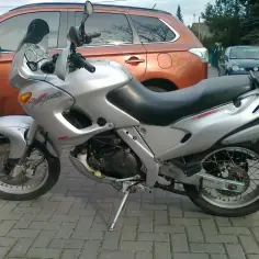 Srebrny motocykl Aprilia Pegaso 650 stoi na bruku obok pomarańczowego SUV-a.