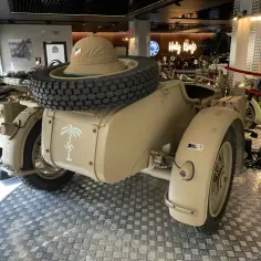 Piaskowy motocykl BMW R75 Sahara z przyczepką, kołem zapasowym i kapeluszem w tle.