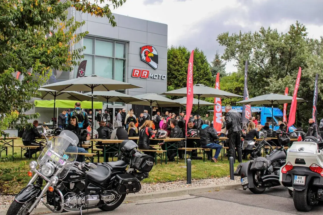 Zjazd motocyklowy przy salonie RRmoto. Ludzie w skórzanych kurtkach siedzą przy stołach pod parasolami, a motocykle stoją zaparkowane.