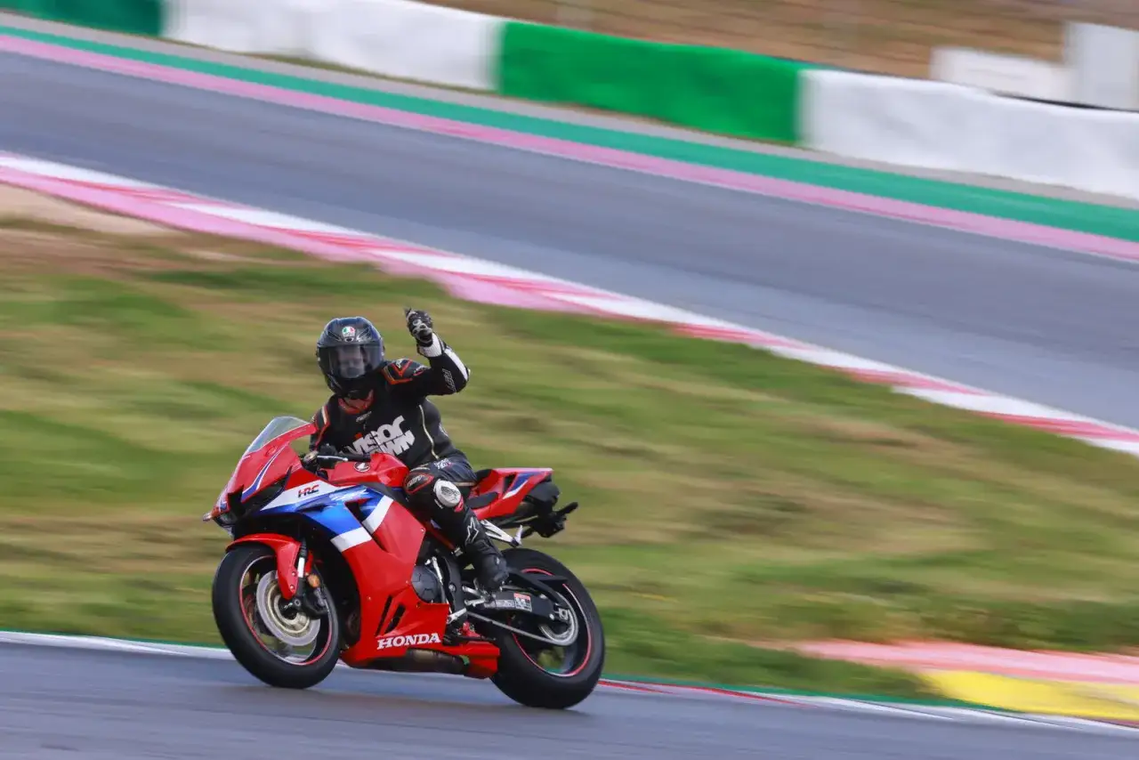 Motocyklista w kombinezonie na torze, pozdrawia ręką, jadąc na czerwonym motocyklu Honda CBR 600RR. Dane techniczne tego modelu robią wrażenie.