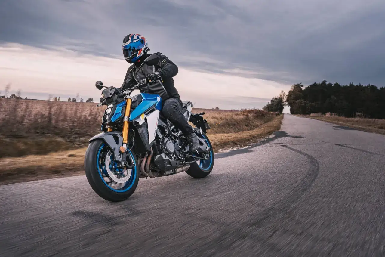 Motocyklista w kasku z flagą Wielkiej Brytanii wykonuje dynamiczny shimmy na niebiesko-białym motocyklu po wiejskiej drodze.