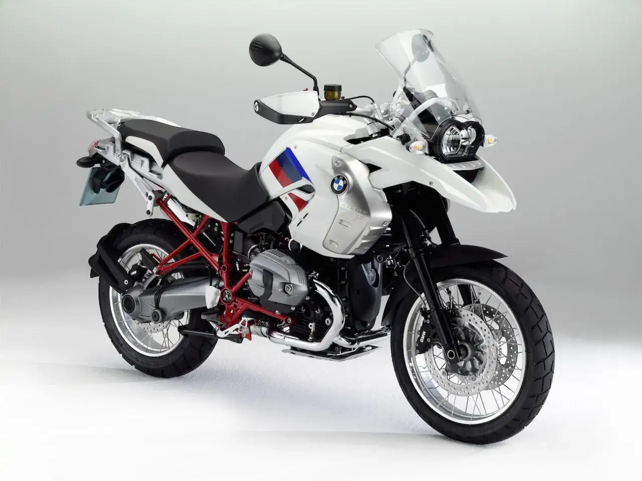 Biały motocykl BMW R1200GS z czerwonym stelażem i flagą na baku. Zwróć uwagę na detale, takie jak opony i zawieszenie.