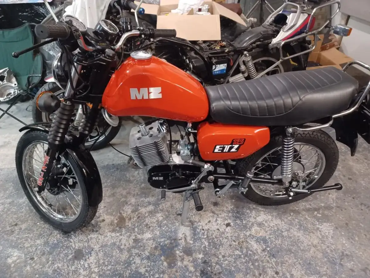 Pomarańczowy motocykl MZ ETZ 125, który wygląda, jakby jego silnik gaśnie na wolnych obrotach, stoi w warsztacie.