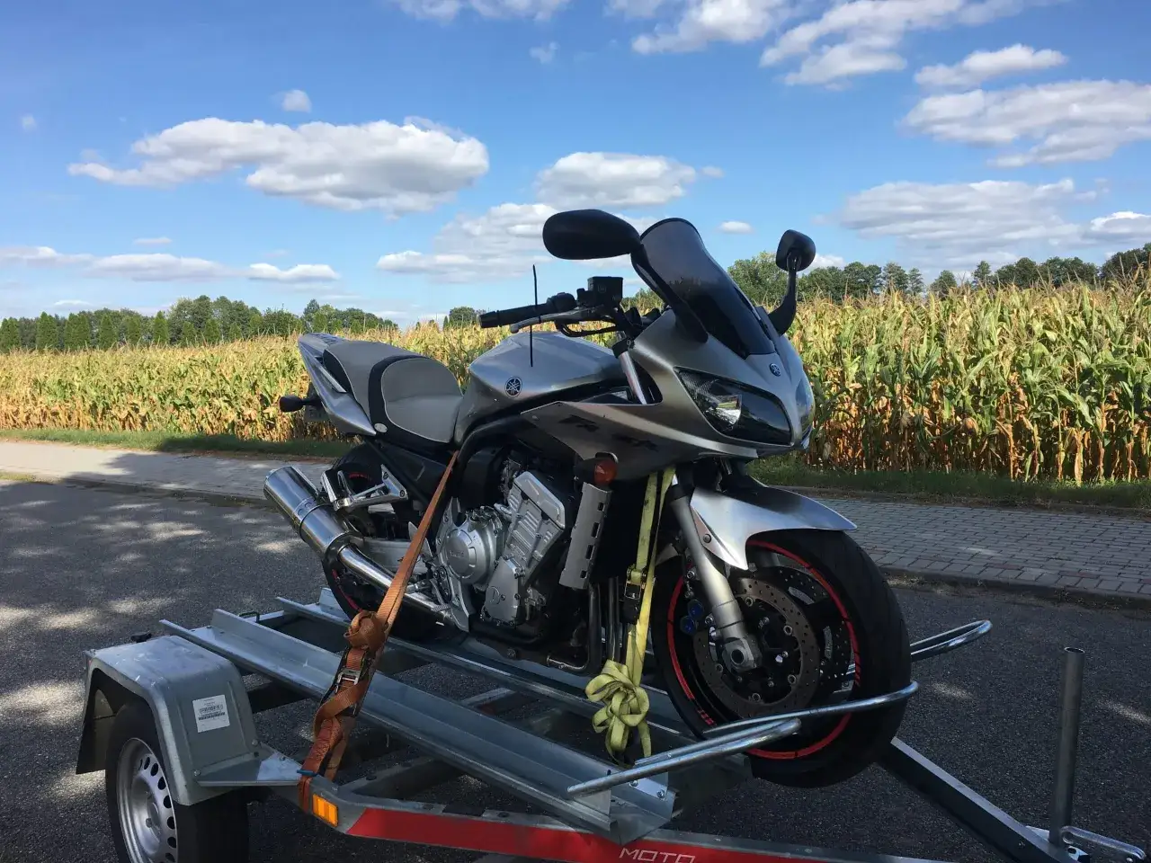Srebrny motocykl Yamaha na przyczepce, gotowy do transportu. W tle pole kukurydzy i błękitne niebo. Czekam na akcyzę za motocykl z Niemiec.
