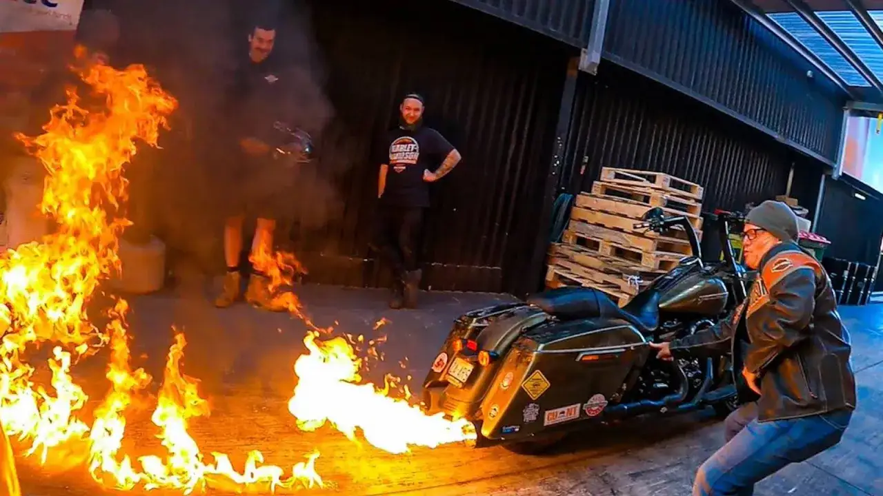 Motocykl Harley-Davidson z potężnym ogniem z wydechu. Dwóch mężczyzn obserwuje widowisko, jeden z nich trzyma w ręku coś, co może być źródłem płomieni.