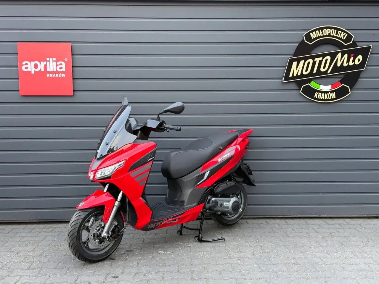 Czerwony skuter Aprilia 1000 SR Max, zaparkowany przed szarym budynkiem z logo salonu MotoMio Kraków.