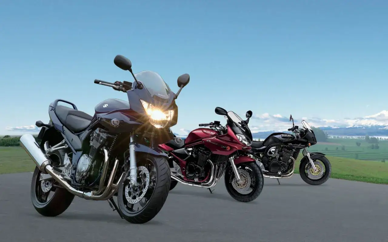 Trzy motocykle Suzuki 1200 w różnych kolorach: granatowym, bordowym i czarnym, stojące na drodze z widokiem na zielone wzgórza.