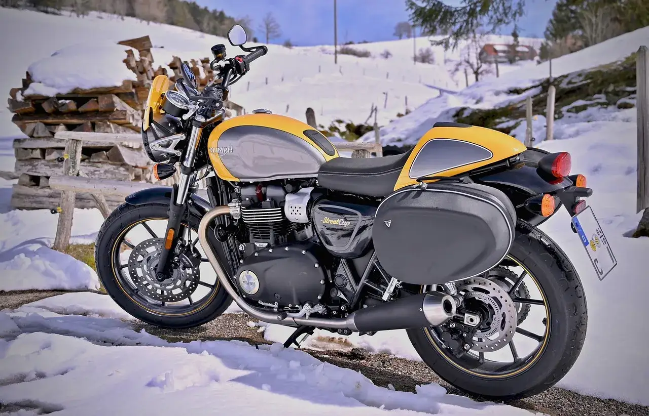 Żółto-szary motocykl Triumph Street Cup w zimowej scenerii. Silnik ma chłodzenie powietrzem, a sakwy boczne dodają mu uroku. Nawet w śniegu czuć jego moc i chęć do jazdy.