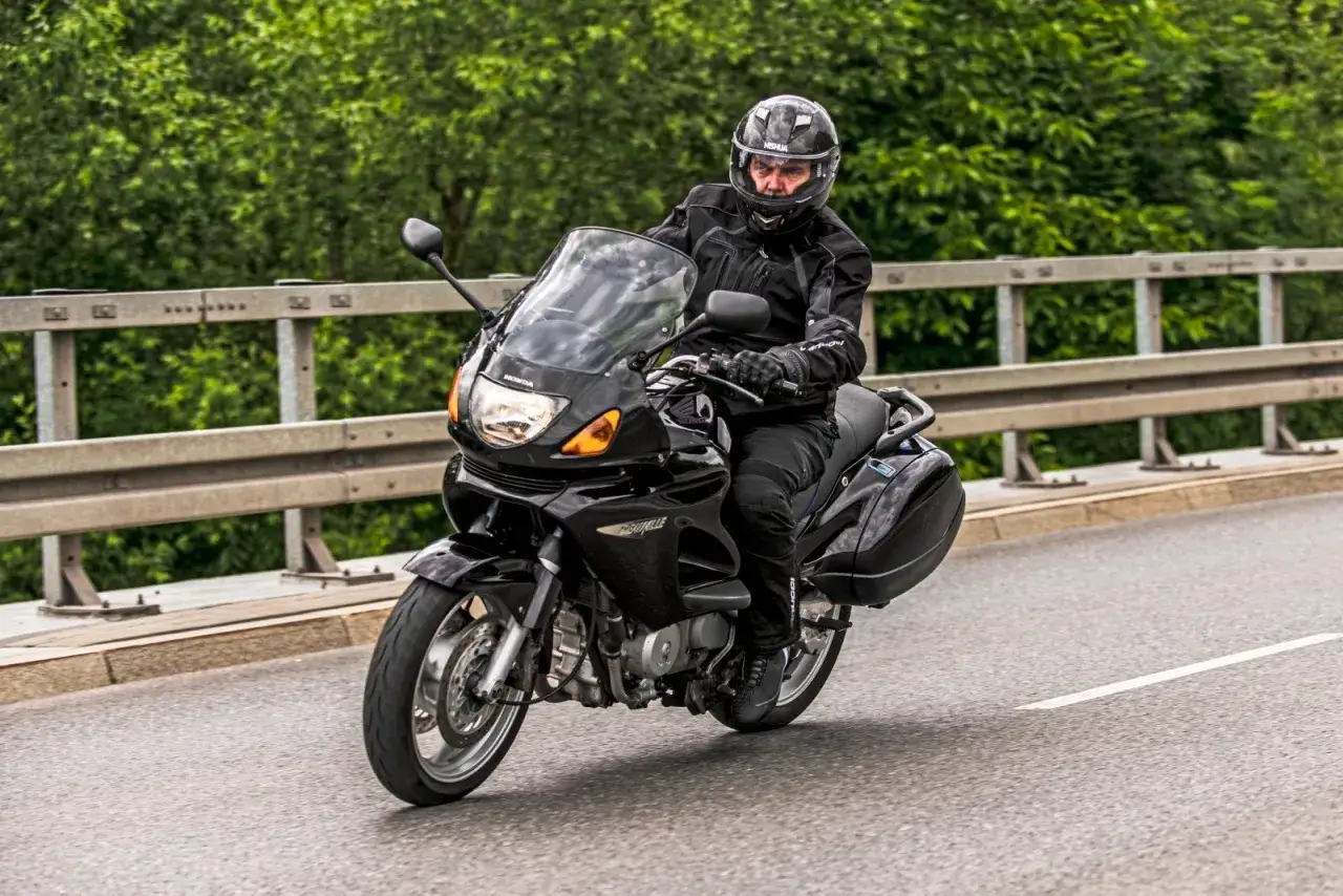 Mężczyzna w kasku i kurtce jedzie na czarnym motocyklu Honda NTV. Maszyna ma zamontowane kufry boczne i dużą szybę.