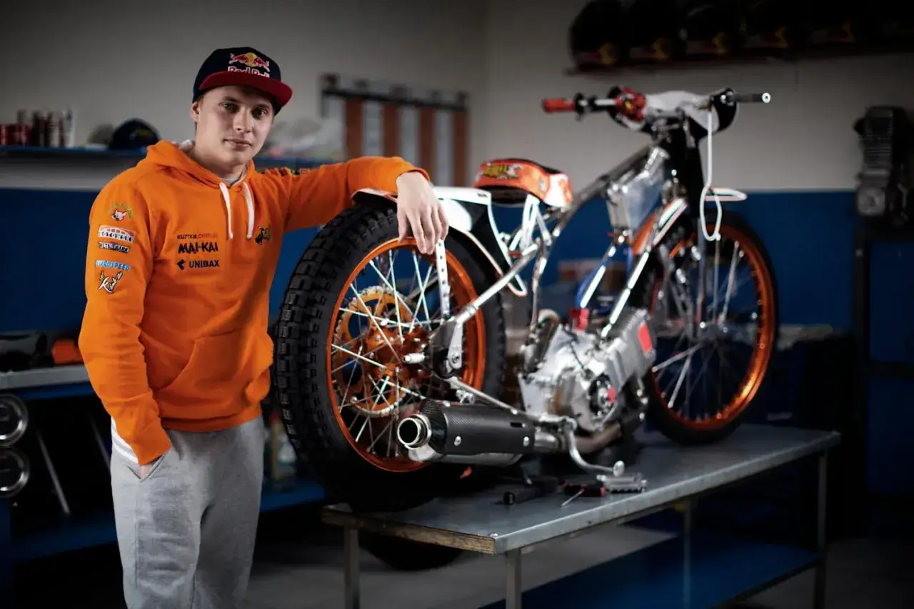 Młody żużlowiec w pomarańczowej bluzie opiera się o swój motocykl żużlowy.