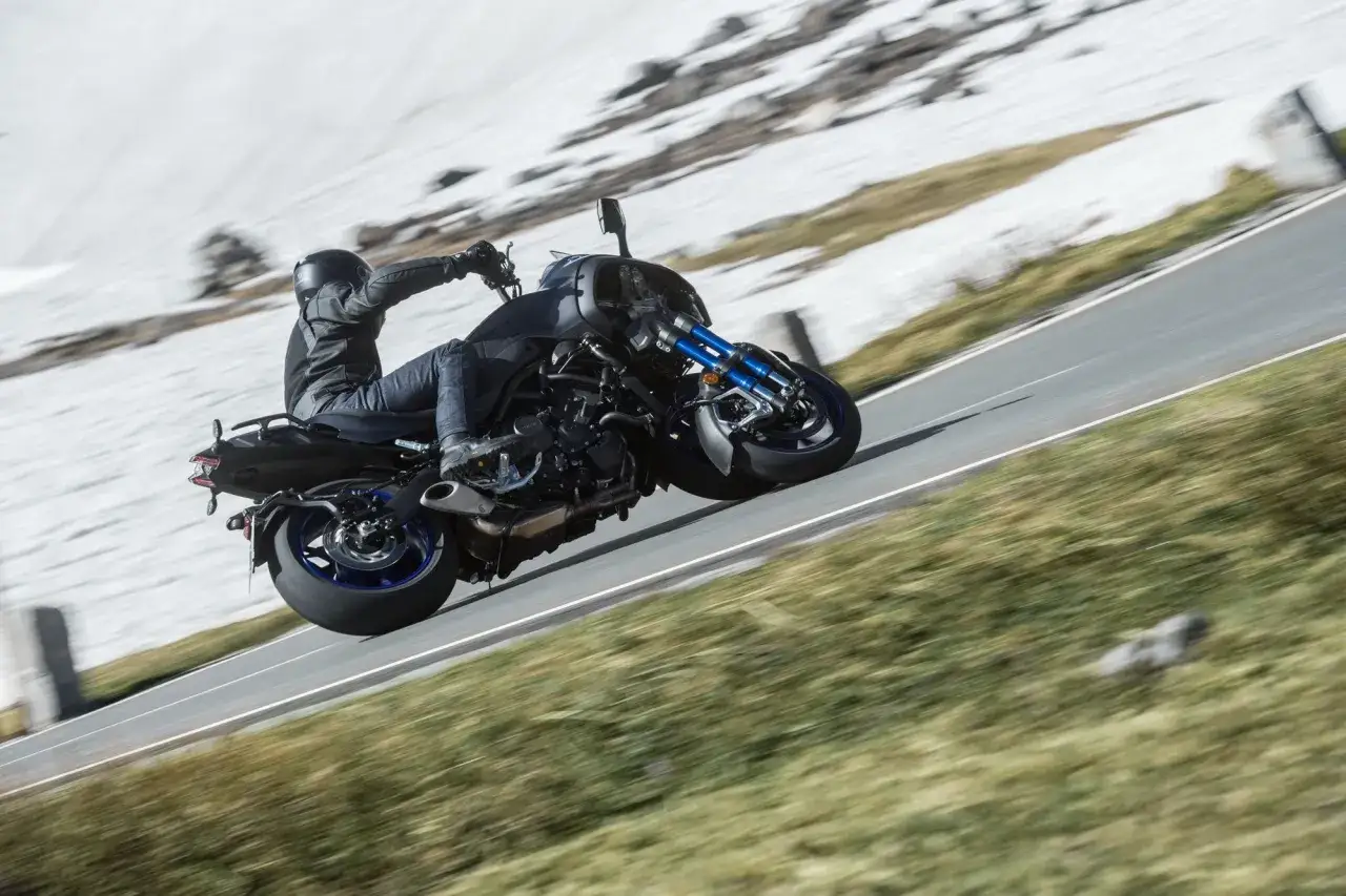 Motocyklista na Yamaha Niken pochyla się na zakręcie górskiej drogi, otoczony wiosenną zielenią i resztkami śniegu.