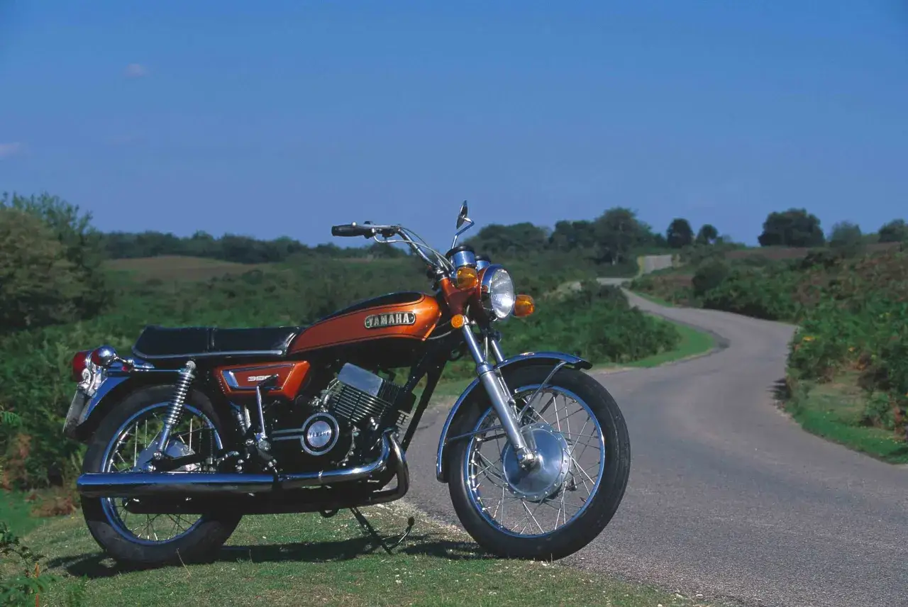 Pomarańczowy motocykl Yamaha RD 350 na tle krętej drogi i zielonych wzgórz.