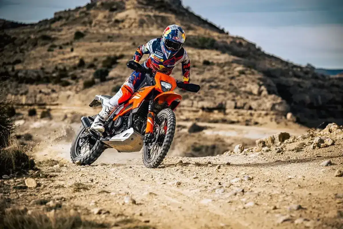 Motocyklista w kasku i stroju crossowym pędzi pomarańczowym motocyklem po pustynnym terenie. Czy na crossa trzeba mieć prawo jazdy? To pytanie zadaje sobie wielu pasjonatów.