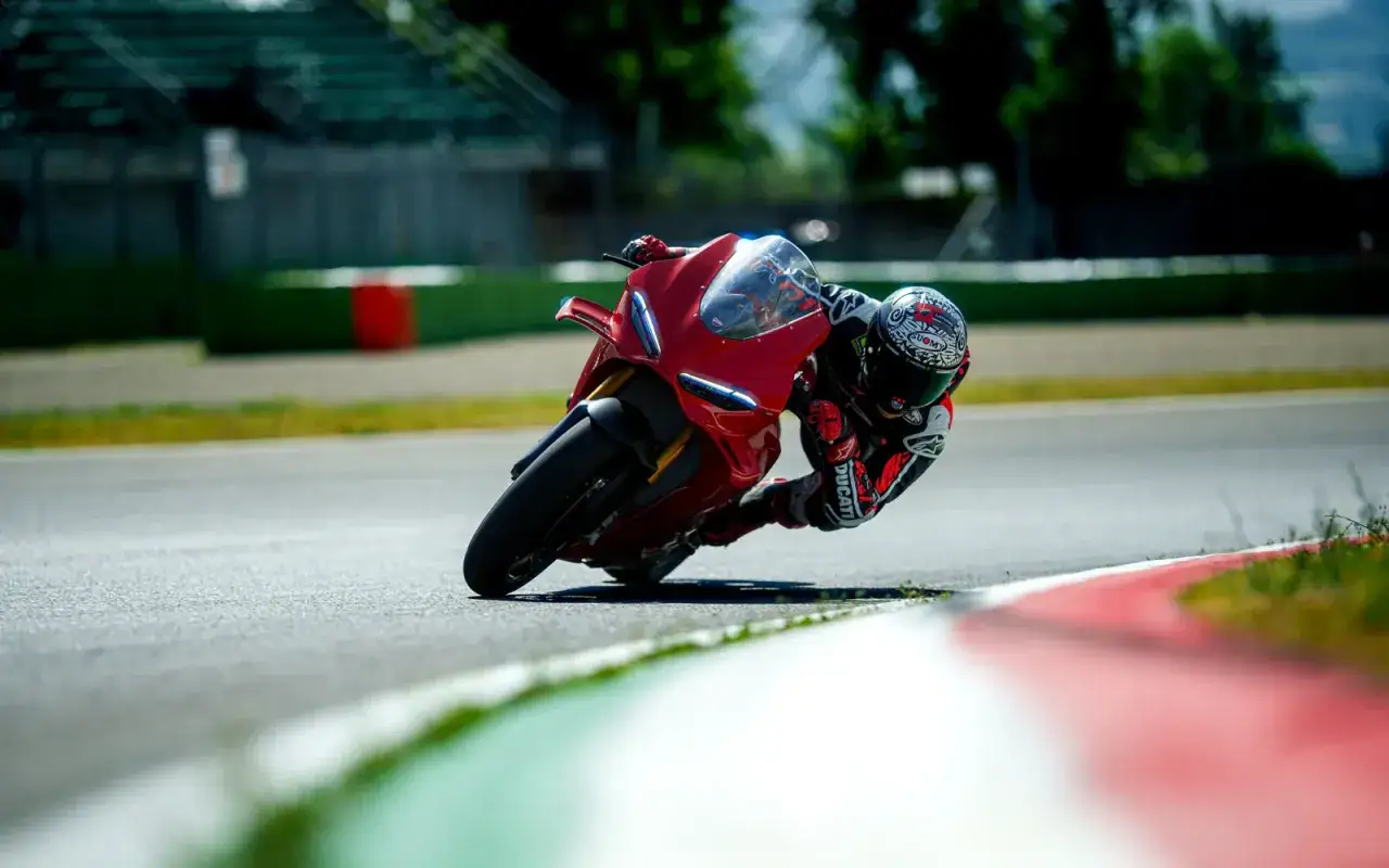 Motocyklista w kasku pochyla się na torze wyścigowym na czerwonym Ducati V4R, z kolanem prawie dotykającym asfaltu.