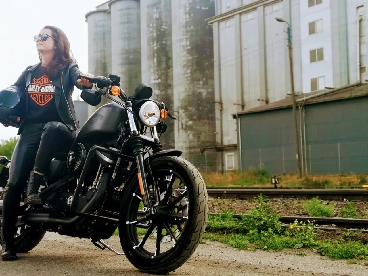 Kobieta w skórzanej kurtce siedzi na czarnym motocyklu Harley-Davidson Sportster 883, gotowa do jazdy obok silosów.