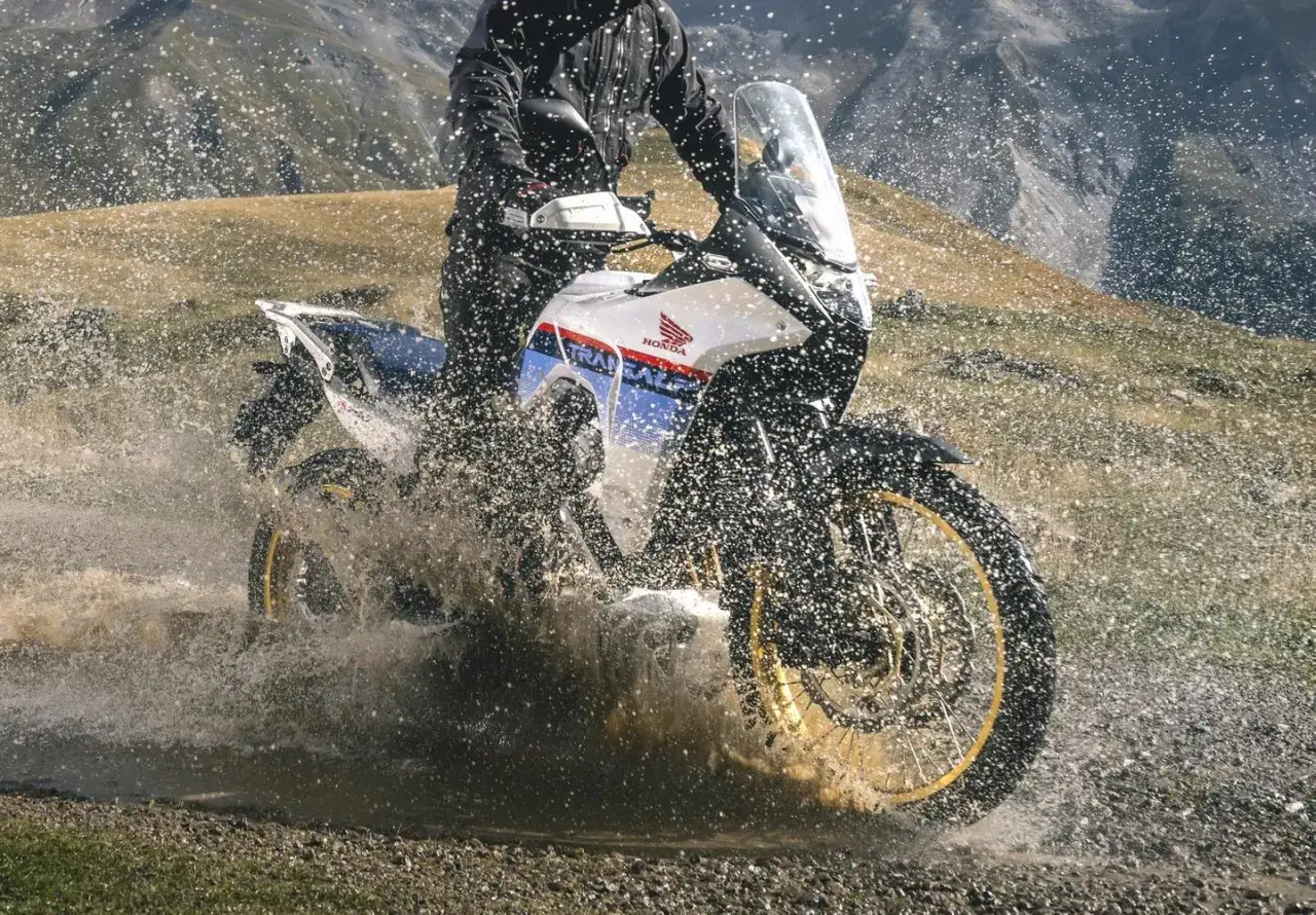 Motocyklista na motocyklu honda turystyk przemierza wodną przeszkodę, wzbijając fontanny wody.